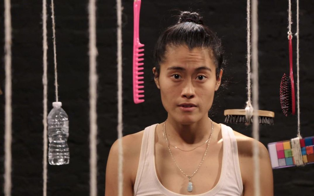 Filmstill aus dem Film "One Big Bag" von Every Ocean Hughes. Man sieht eine Person vor einem schwarzen Hintergrund, um sie herum hängen Gegenstände an Schnüren.