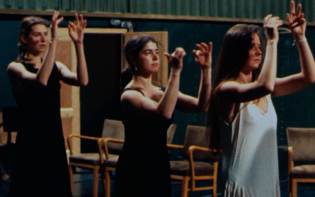 Filmstill aus Was tun Pina Bausch und ihre Tänzer in Wuppertal?: Drei Frauen stehen hintereinander auf einer Bühne und haben gehobene Arme.