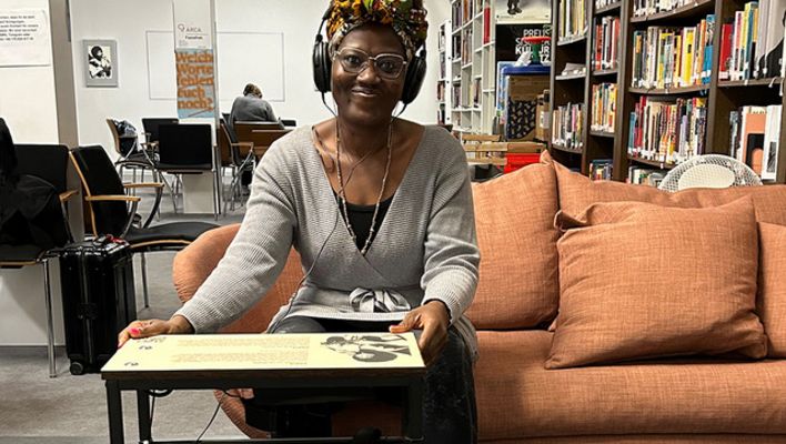 A woman is sitting on a sofa in a library, wearing headphones and smiling at the camera. In front of her is a low table covered with text and images.