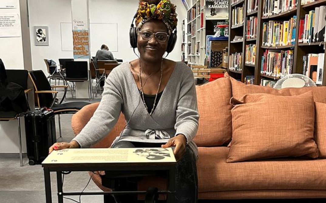 A woman is sitting on a sofa in a library, wearing headphones and smiling at the camera. In front of her is a low table covered with text and images.