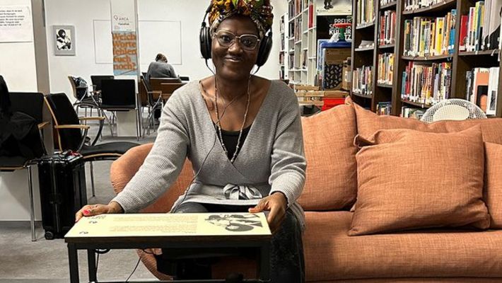 Eine Frau sitzt in einer Bibliothek auf einem Sofa, trägt Kopfhörer und blickt lächelnd in die Kamera. Vor ihr steht ein tiefer Tisch, der mit Text und Bildern bedruckt ist.