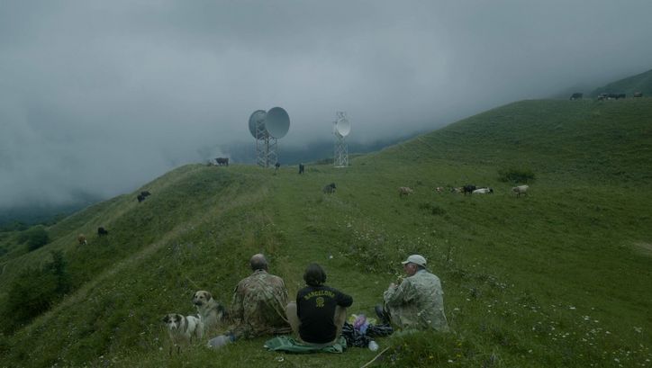 Film still from THE HARVEST. Three men are seen sitting on the grass on a hill. Cows are grazing in the background and two radio towers with satellite dishes can be seen.
