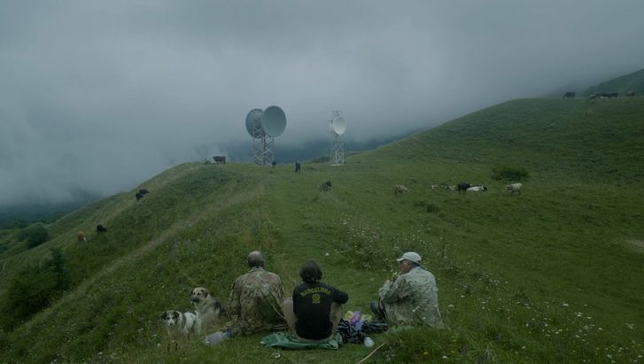 Film still from THE HARVEST. Three men are seen sitting on the grass on a hill. Cows are grazing in the background and two radio towers with satellite dishes can be seen.