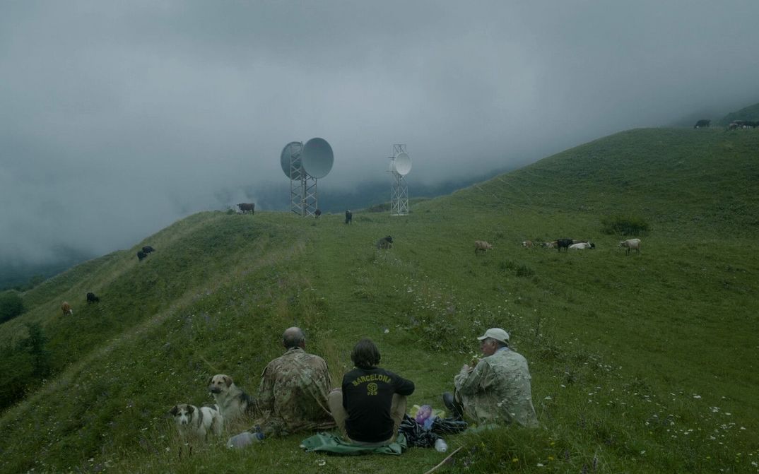 THE HARVEST Film still from THE HARVEST. Three men are seen sitting on the grass on a hill. Cows are grazing in the background and two radio towers with satellite dishes can be seen.