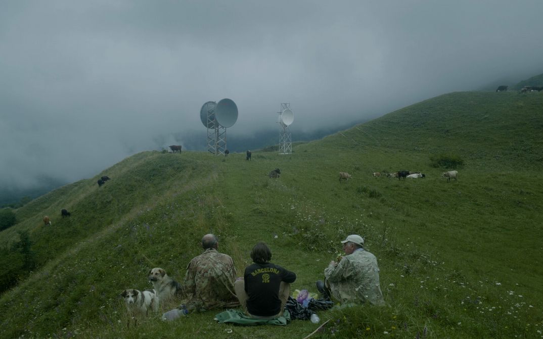 THE HARVEST Filmstill aus THE HARVEST. Auf einem Hügel sieht man drei Männer im Gras sitzen. Im Hintergrund grasen Kühe und sind zwei Funktürme mit Satellitenschüsseln zu sehen.