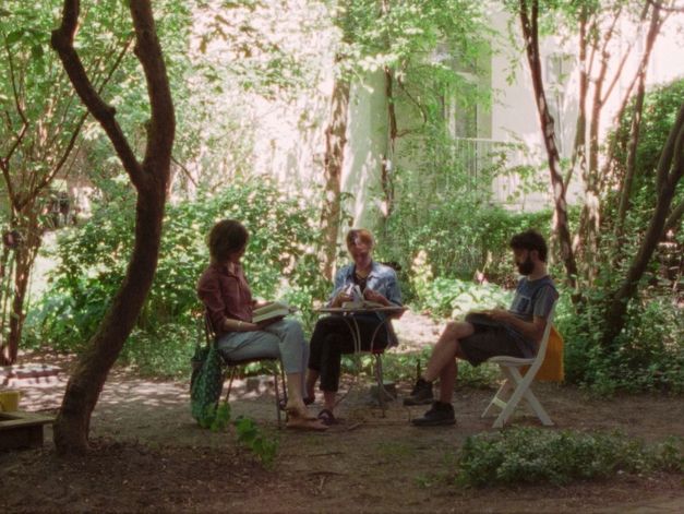 Drei Personen sitzen an einem Tisch im Garten und lesen und sprechen, umgeben von Bäumen und dichtem Grün.