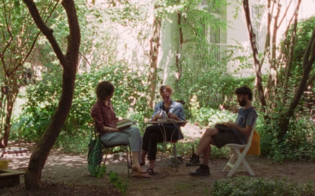Drei Personen sitzen an einem Tisch im Garten und lesen und sprechen, umgeben von Bäumen und dichtem Grün.