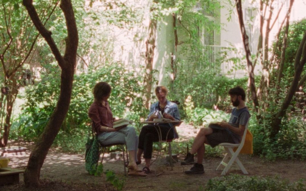 Drei Personen sitzen an einem Tisch im Garten und lesen und sprechen, umgeben von Bäumen und dichtem Grün.