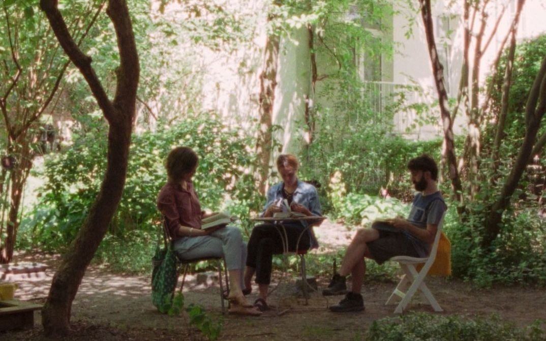 Three people sit at a table in a garden reading and talking, surrounded by trees and dense greenery.