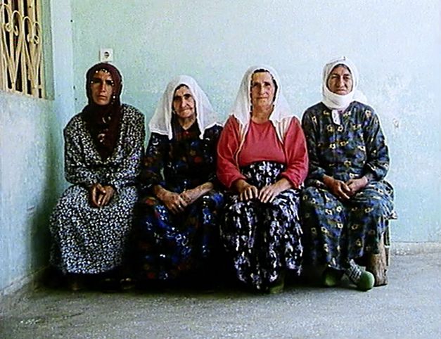 Still from the film „Mein Vater, der Gastarbeiter“ by Yüksel Yavuz. Four women in headscarves sit next to each other on a bench against the wall of a house.