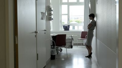 Film still from PALLIATIVSTATION (Palliative Care Unit) by Philipp Döring. The image shows a woman leaning against the wall in a hospital room.