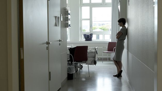 Film still from PALLIATIVSTATION (Palliative Care Unit) by Philipp Döring. The image shows a woman leaning against the wall in a hospital room.