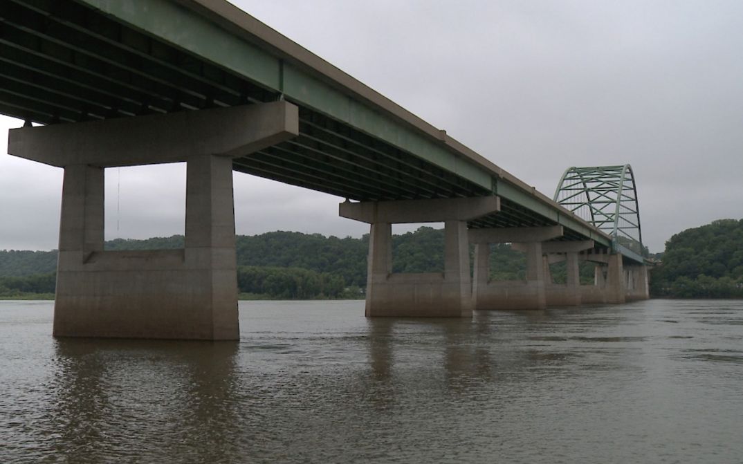 Große Betonbrücke überspannt breiten Fluss, bewaldete Hügel im Hintergrund.