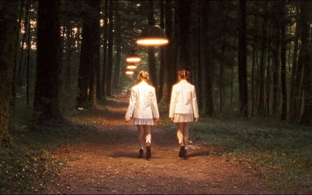 Film still from INNOCENCE: Two girls with braids and school uniforms are walking along a forest path; you see them from behind. Hanging lamps light up the path.