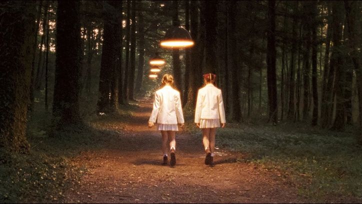Film still from INNOCENCE: Two girls with braids and school uniforms are walking along a forest path; you see them from behind. Hanging lamps light up the path.