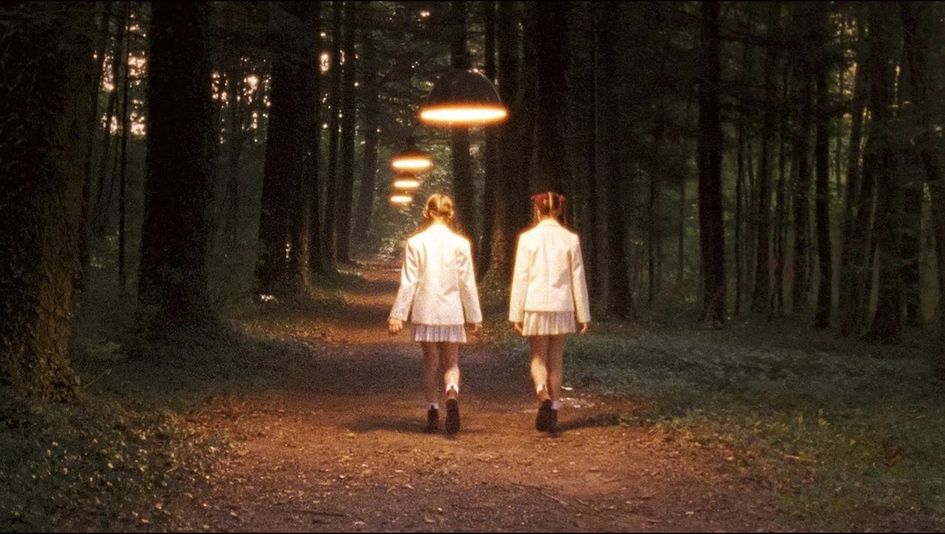 Filmstill aus INNOCENCE: Zwei Mädchen mit Zöpfen und Schuluniform gehen einen Waldweg entlang, man sieht sie von hinten. Aufgehängte Lampen erleuchten den Weg.