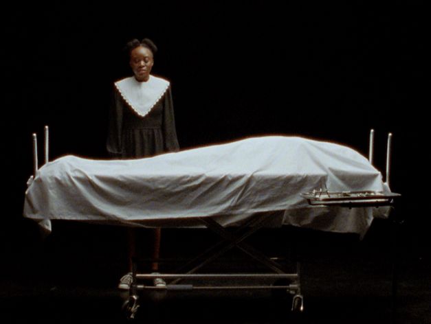 Still from the film "Cette Maison (This House)" by Miryam Charles. A teenage girl dressed in black and white stands behind a body covered by a sheet on a medical bed, in a black soundstage.