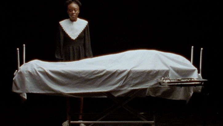 Still from the film "Cette Maison (This House)" by Miryam Charles. A teenage girl dressed in black and white stands behind a body covered by a sheet on a medical bed, in a black soundstage.