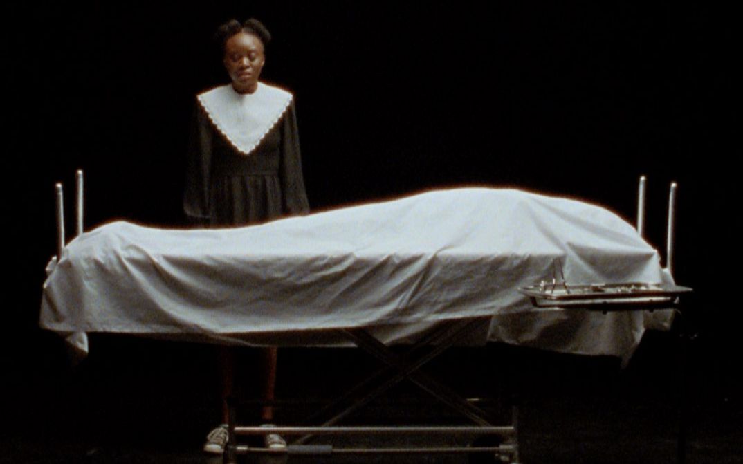 Filmstill aus „This House“ von Miryam Charles. Auf einer dunklen Bühne steht eine junge Frau in Trauerkleidung vor einer Krankenhausliege, auf der von einem weißen Laken zugedeckt ein Körper liegt.