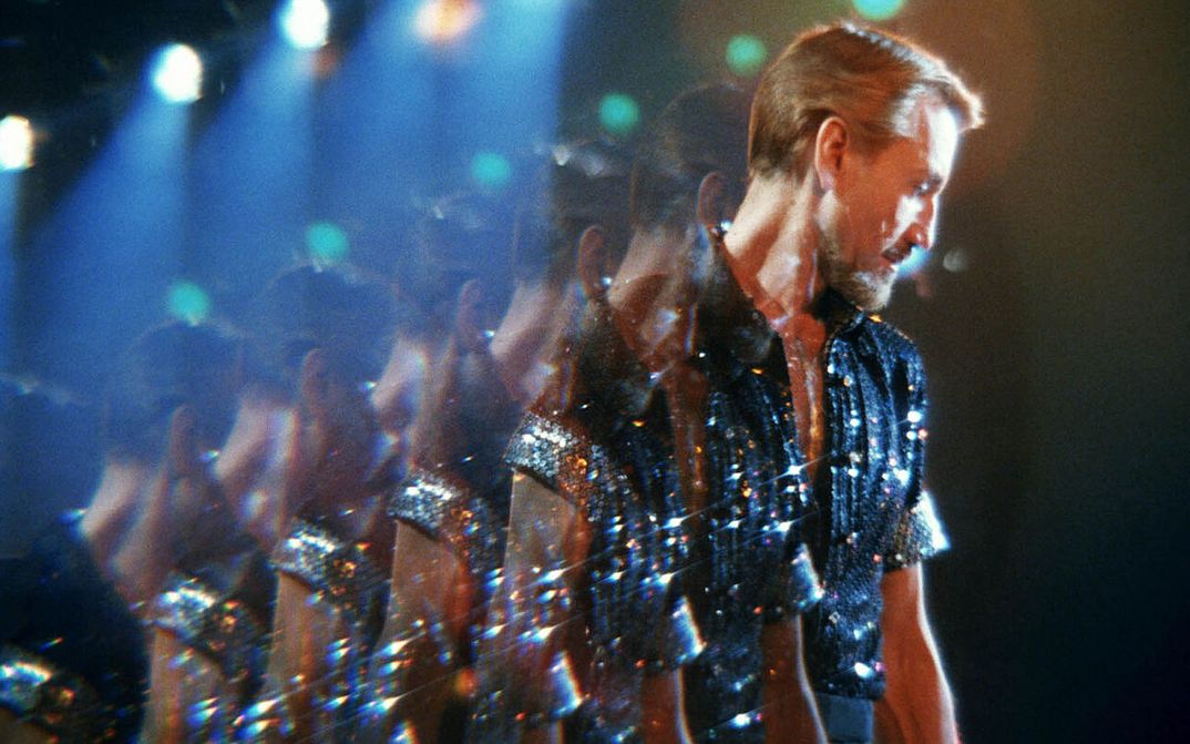 Filmstill aus ALL THAT JAZZ: Ein Mann mit blonden Haaren und einem blau glitzerndem Paillettenhemd steht im Diskolicht. Durch eine Spiegelung erscheint er mehrfach kopiert.
