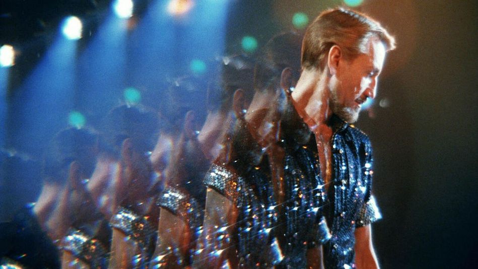 Filmstill aus ALL THAT JAZZ: Ein Mann mit blonden Haaren und einem blau glitzerndem Paillettenhemd steht im Diskolicht. Durch eine Spiegelung erscheint er mehrfach kopiert.