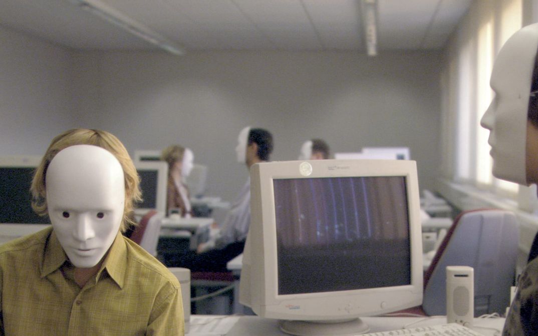 Filmstill aus FALSCHER BEKENNER: Einige Personen sitzen in einem Großraumbüro vor Computern. Alle tragen weiße Masken, die das ganze Gesicht bedecken.