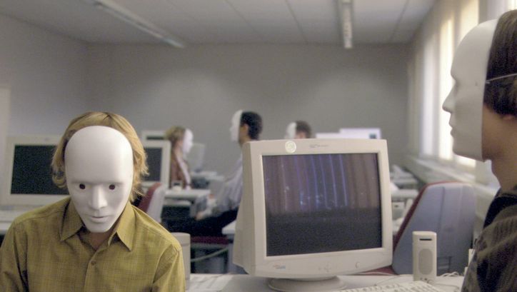 Filmstill aus FALSCHER BEKENNER: Einige Personen sitzen in einem Großraumbüro vor Computern. Alle tragen weiße Masken, die das ganze Gesicht bedecken.