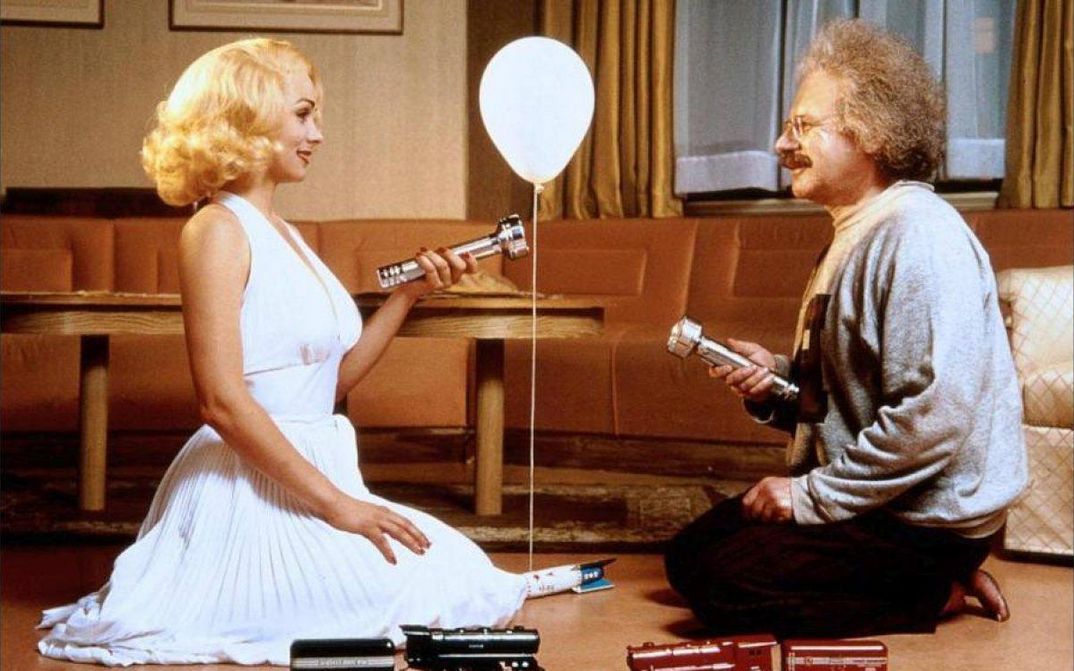 Film still from INSIGNIFICANCE: Two people, portraying Marilyn Monroe and Albert Einstein, are kneeling facing each other in a hotel room. They are shining flashlights on each other, and a balloon is floating between them.