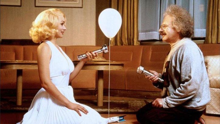Film still from INSIGNIFICANCE: Two people, portraying Marilyn Monroe and Albert Einstein, are kneeling facing each other in a hotel room. They are shining flashlights on each other, and a balloon is floating between them.