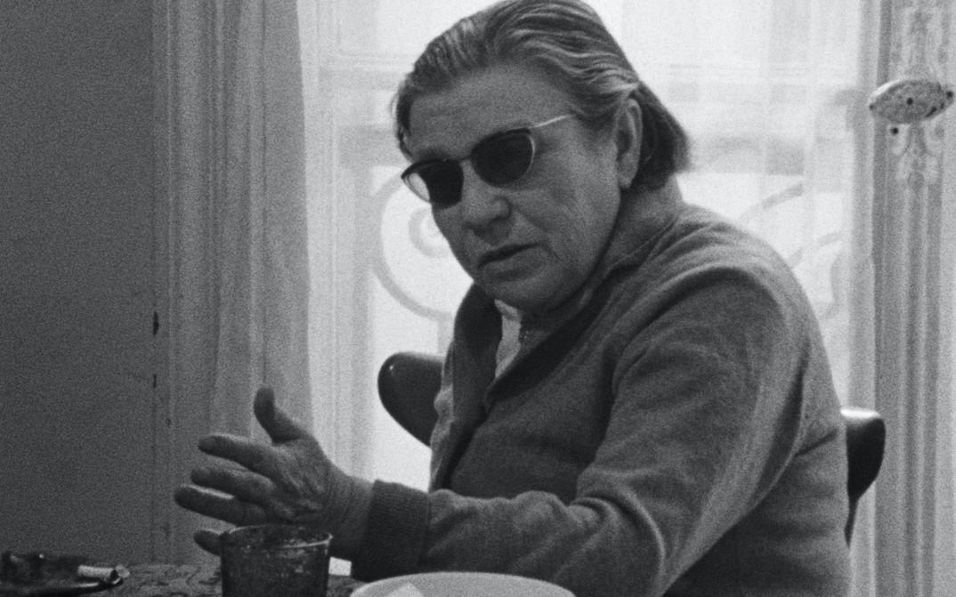 Filmstill aus NUMÉRO ZÉRO: Eine alte Frau mit dunkel getönter Brille sitzt gestikulierend an einem Tisch.