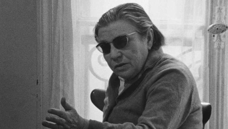 Filmstill aus NUMÉRO ZÉRO: Eine alte Frau mit dunkel getönter Brille sitzt gestikulierend an einem Tisch.