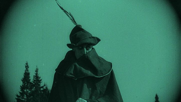 Filmstill aus NOSFERATU: Monochrom grün eingefärbtes Bild eines Mannes mit Mantel und Hut auf einem Pferd.