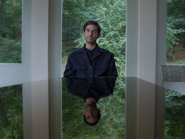 Still from „De Facto" von Selma Doborac. A man sitting at a black table, which mirrors his face and torso. Behind him you can see bushes and trees.
