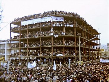 Still from the film "Între revoluții" by Vlad Petri. Masses of people are huddled across four stories in a building structure. There are even more people on the street in front of the structure. There is a fabric poster hanging off the building, with red and black writing in Farsi.