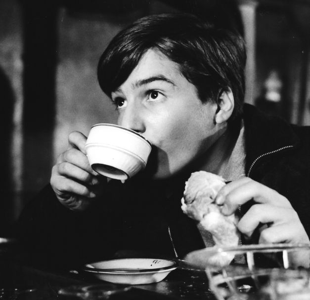 Film still from BOULEVARD: A boy is sitting at a table, drinking a coffee and holding a croissant.