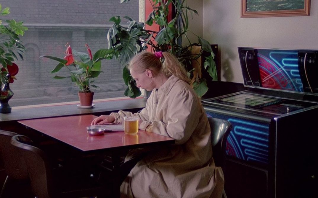 Film still from THE MATCH FACTORY GIRL: A young woman sits alone at an inn table reading a magazine.