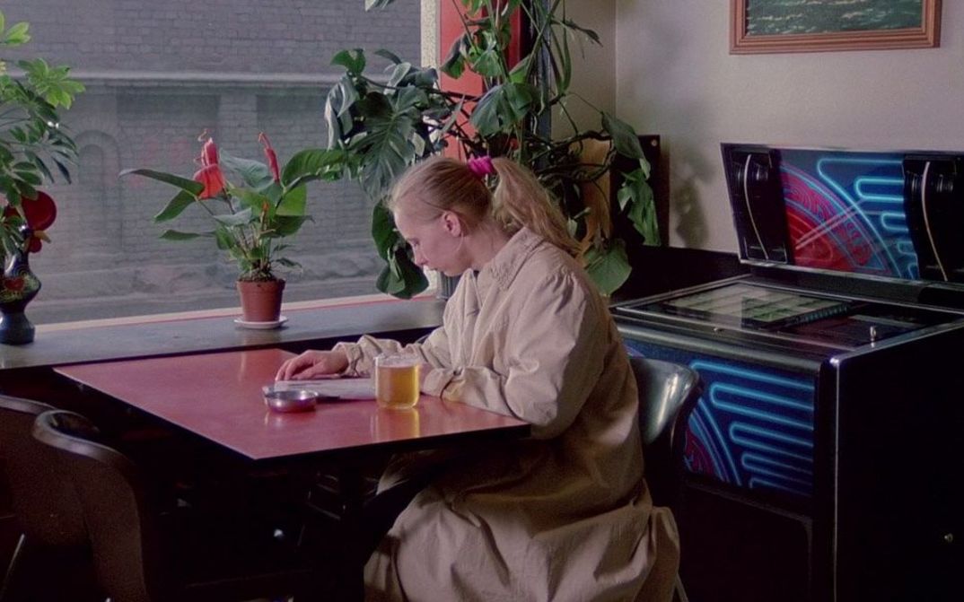 Filmstill aus DAS MÄDCHEN AUS DER STREICHHOLZFABRIK: Eine junge Frau sitzt allein an einem Wirtshaustisch und liest in einer Zeitschrift.