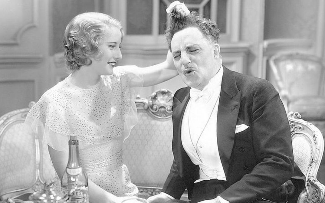 Film still from BABY FACE: A young woman and an older man, both elegantly dressed, sit next to each other on the sofa. She playfully tugs his hair.