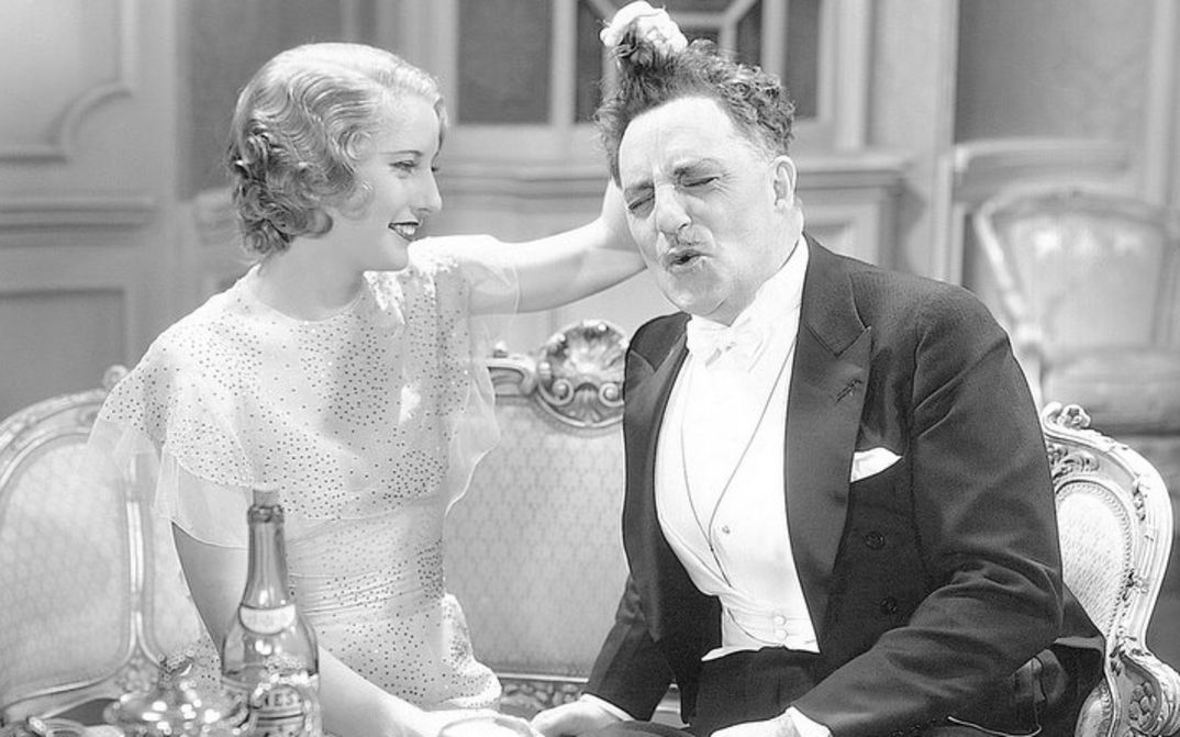 Film still from BABY FACE: A young woman and an older man, both elegantly dressed, sit next to each other on the sofa. She playfully tugs his hair.