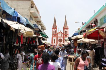 Die koloniale Bauweise der Herz-Jesu Kathedrale hebt sich deutlich im Hintrgrund eines geschäftigen Markts in einer Straße der togoischen Hauptstadt Lomé ab.