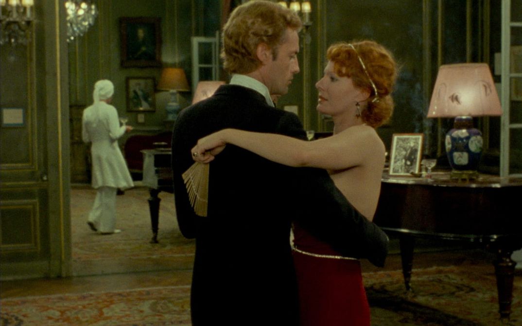 Filmstill from INDIA SONG: A man and a woman in evening dress dance together. A servant can be seen in the background. The rooms appear sumptuous.