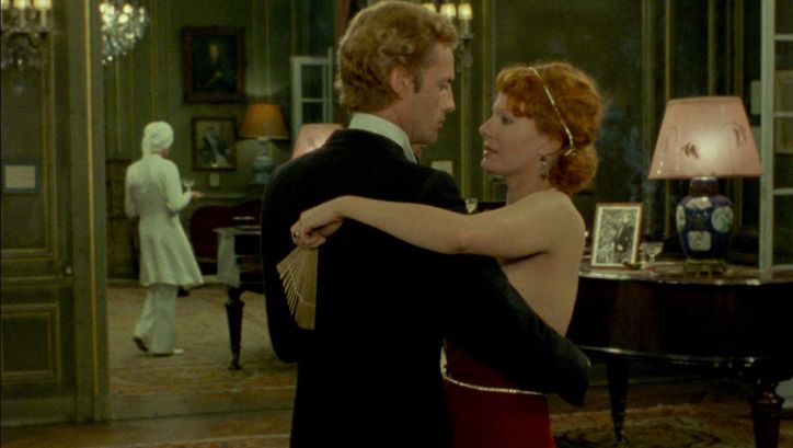 Filmstill aus INDIA SONG: Ein Mann und eine Faru in Abendgarderobe tanzen gemeinsam. Im Hintergrund ist eine Bedienstete zu sehen. Die Räume wirken prunkvoll.