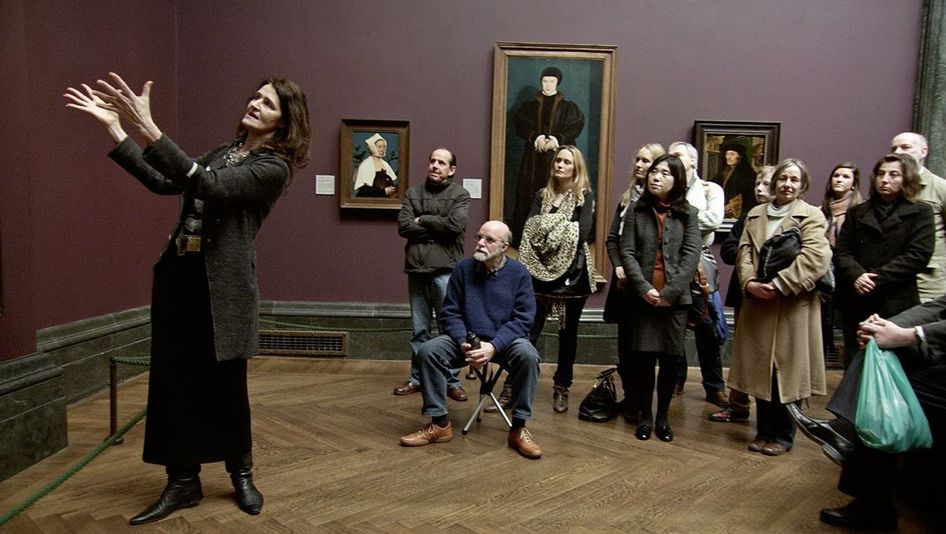 Film still from NATIONAL GALLERY. A woman explains a work of art to a group of museum visitors.