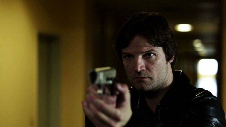 Filmstill from IM SCHATTEN: A man points a gun at something or someone. He is standing in a corridor.
