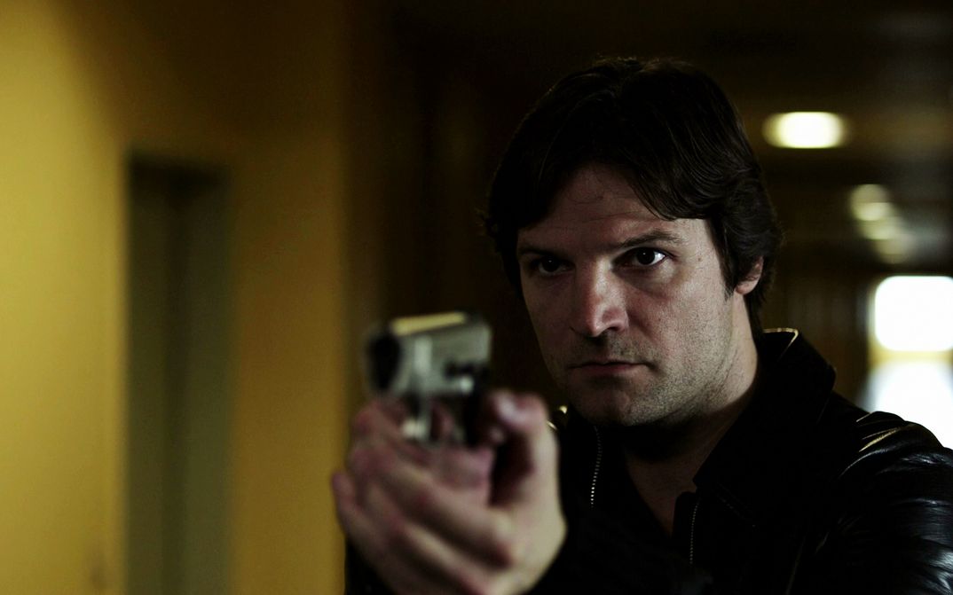 Filmstill from IM SCHATTEN: A man points a gun at something or someone. He is standing in a corridor.