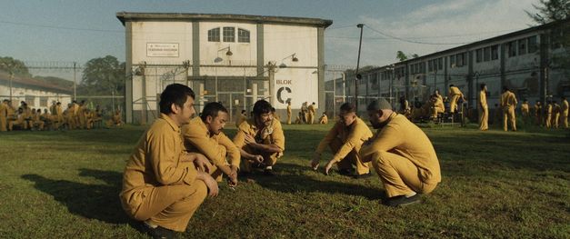 Mehrere Männer in gelber Kleidung hocken auf einer Wiese, offensichtlich ein Gefängnishof, im Hintergrund ein Gefängnisgebäude.