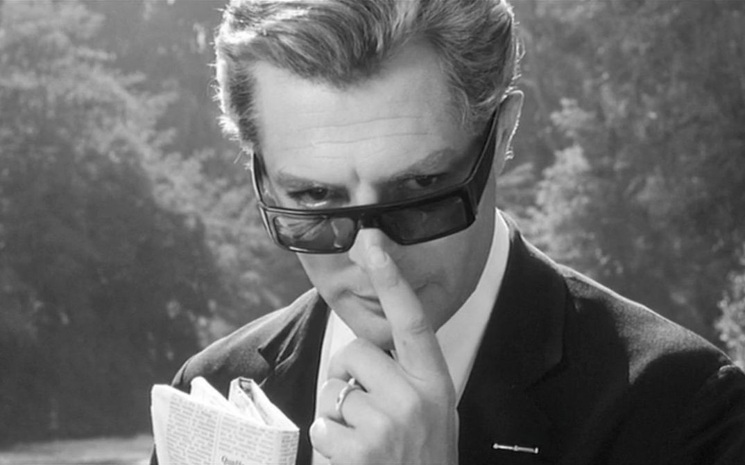 Filmstill aus OTTO E MEZZO: Marcello Mastroianni in einem eleganten Anzug, er blickt über seine Sonnenbrille direkt in die Kamera.
