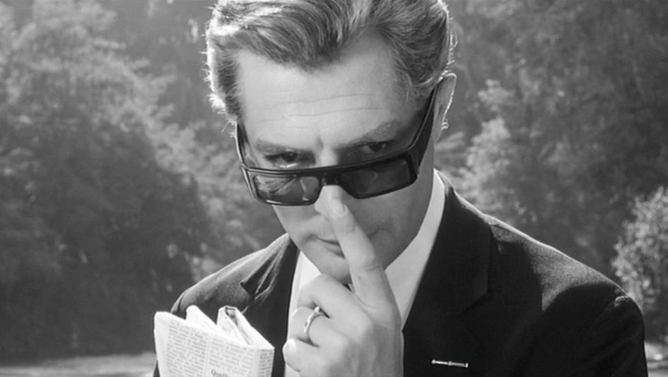 Filmstill aus OTTO E MEZZO: Marcello Mastroianni in einem eleganten Anzug, er blickt über seine Sonnenbrille direkt in die Kamera.