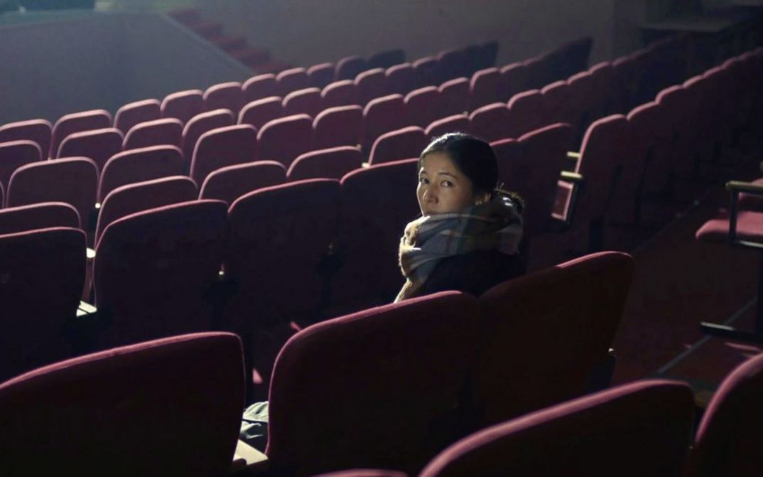 Filmstill aus „Akyn (Poet)“ von Darezhan Omirbayev. Eine Frau sitzt alleine in einem leeren Theatersaal. Sie dreht sich um und schaut in die Kamera.