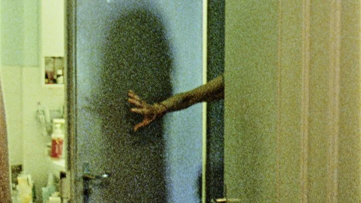 Film still from "Spuren von Bewegung vor dem Eis" by René Frölke. It shows a bathroom. The bathroom door is held open by an arm. 
